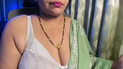 Media: Video of a South Asian woman with medium brown skin, wearing a white lace bra, green dupatta, and gold necklace, against patterned curtains in a dimly lit room.