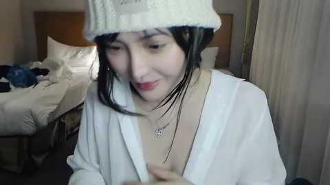 Video of an East Asian woman with wet, dark hair, wearing a white knit beanie and robe, sitting on a bed in a dimly lit room.