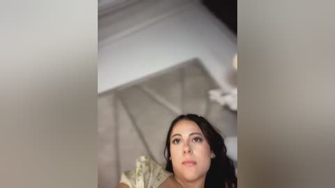 Video of a young woman with long, dark hair, light skin, and subtle makeup, wearing a white dress, kneeling on a beige carpet, looking upward. The background features a white table and blurred white fabric.