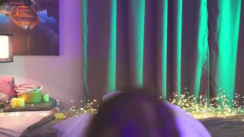 Video of a dark-haired person, blurred, lying on a bed with a teal-green curtain backdrop, fairy lights, and a colorful wooden boat model on a table.