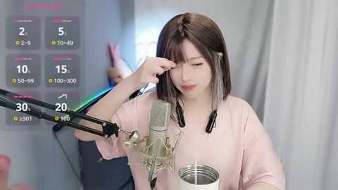 Video of a young Asian woman with straight brown hair, wearing a light pink t-shirt, recording herself singing into a microphone. Background features a gray curtain and a digital display showing song progress.