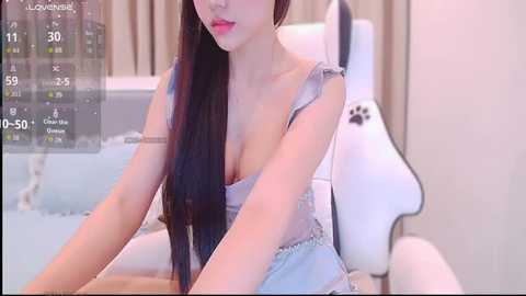 Video of an East Asian woman with long black hair, wearing a revealing light blue top, sitting on a white gaming chair. Background includes a computer screen displaying a game.