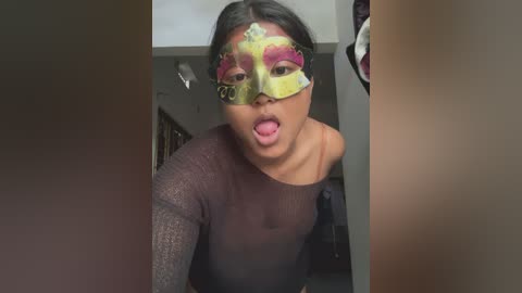 Video of a young woman with medium brown skin, wearing a glittery black top, sticking out her tongue and sporting a green and gold masquerade mask with pink eyeshadow. She's indoors, in a dimly lit room with blurred background.