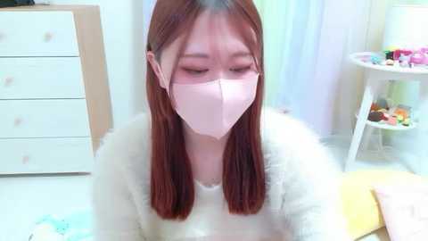 Media: A video of a young Asian woman with long brown hair, wearing a pink face mask, sitting indoors. She's in a cozy, bright room with light-colored furniture and a toy shelf in the background.