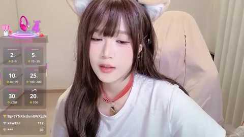 Video of a young East Asian woman with fair skin, long dark hair, and cat ears, wearing a white t-shirt and red choker. She has a neutral expression, sitting indoors.