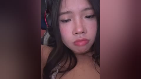 Video of an Asian woman with long black hair, wearing a headset, and light makeup. She has a neutral expression and is wearing a tan top.