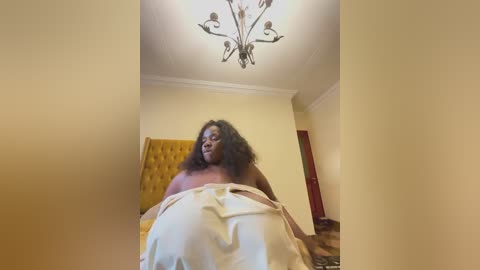 A video of a nude, dark-skinned woman with curly hair, covered in a white sheet, lying on a bed with a yellow quilt. The room features a chandelier and beige walls.