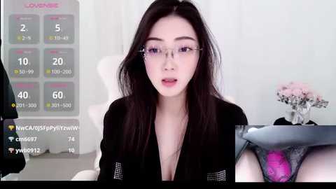 Video of a young Asian woman with long black hair, wearing glasses, black jacket, and a low-cut top, seated in a white chair, in a modern room with a calendar and flowers in the background.