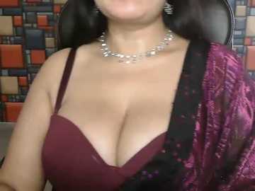 Video of a woman with light skin and long dark hair, wearing a low-cut burgundy bra and a black and purple floral-patterned shawl, against a mosaic-tiled wall. She accessorizes with a silver necklace.