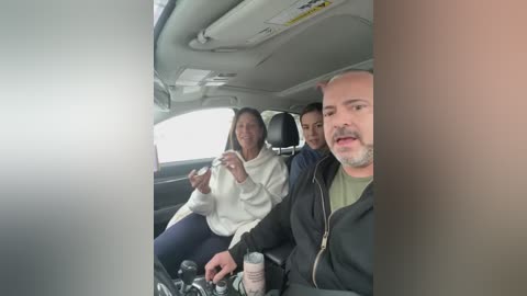 Video of a smiling couple in a taxi, the woman in a white hoodie and jeans, the man in a black jacket, and another man in the backseat.