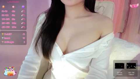 A video of a light-skinned Asian woman with long black hair, wearing a white off-shoulder top that reveals cleavage, sitting on a beige couch in a softly lit room.