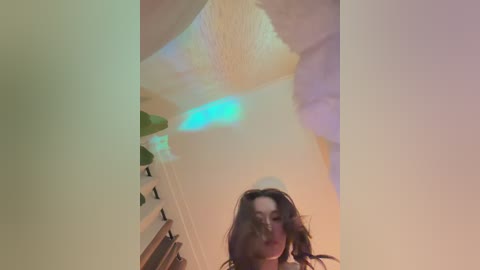 A blurry video of a woman with long brown hair, wearing a white sweater, standing in a dimly lit room with a green plant and a blue light.