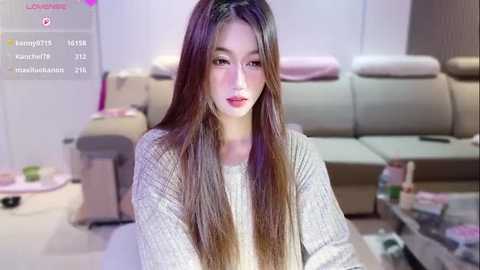 Video of a young East Asian woman with long, straight brown hair, wearing a grey sweater, sitting on a couch in a modern living room with a TV and glass table.