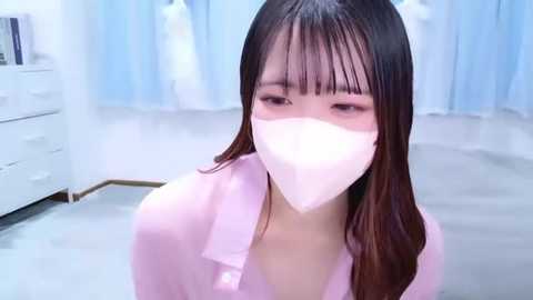 A video of a young East Asian woman with long, straight, dark hair, wearing a pink shirt and a white face mask, sitting in a sterile, white-walled room with light blue curtains.