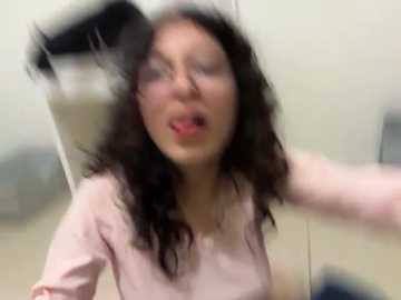 Video of a woman with long black hair and a pink shirt, sitting with her eyes closed and mouth open in a playful, possibly surprised expression. Background is blurry, possibly indoors.
