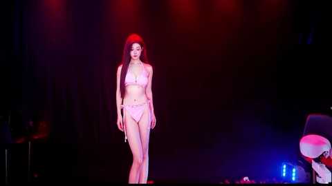Video of an Asian woman with long black hair in a red headband, wearing a pink bikini, walking confidently on a stage under dim red and blue lights.