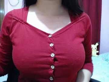 Video of a woman with medium skin tone wearing a red button-up cardigan with a white undershirt, standing in a room with a blue bedspread, red curtains, and a Pikachu plush toy.