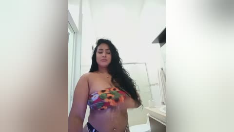 Video of a Latina woman with long black hair in a colorful, patterned strapless top, standing in a bathroom with white walls, a shower curtain, and towels.