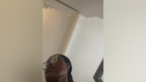 Video of a young, dark-skinned man with glasses and braided hair, captured from a low angle, showing his face and chest. He stands in a dimly lit room with cream-colored walls.