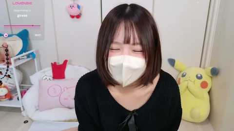 Video of an Asian woman with straight black hair, wearing a white face mask, black top, and Pikachu plush toys in a white room.