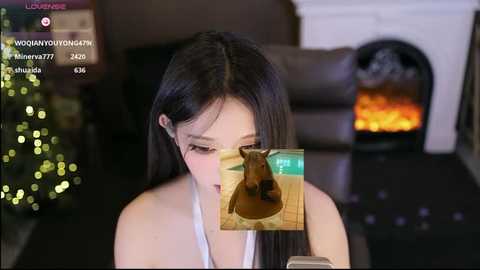 A video of a young Asian woman with long black hair, wearing glasses and a white top, holding a smartphone. Superimposed on the image is a virtual dog. In the background, a lit Christmas tree and a fireplace are visible.