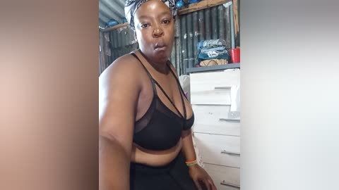 Video of a plus-sized Black woman with medium brown skin, wearing a black bra and pants, standing in a cluttered room with a white dresser, clothes, and a red cup.