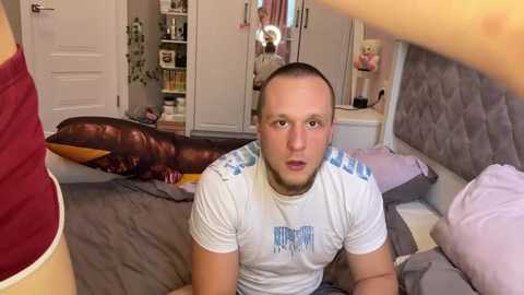 A video of a bald, muscular man in a white T-shirt, sitting on a gray bed with a stuffed shark, in a cluttered bedroom with a mirror.