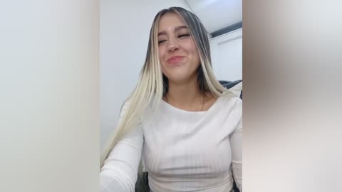 Video of a young woman with long, straight, blonde hair, fair skin, and a white ribbed long-sleeve top, smiling with her eyes closed, taken indoors with a blurred background.