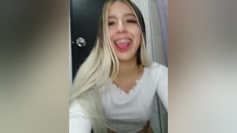 Video of a young, blonde, Caucasian woman with a playful expression, sticking out her tongue, wearing a white long-sleeve top, taken indoors.