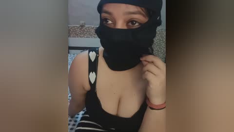 Video of a woman with medium skin tone, wearing a black face mask, black beanie, and a low-cut black top revealing cleavage, in a dimly lit room with tiled walls.