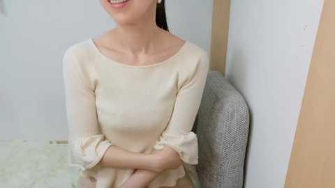 Video of a smiling Asian woman with long black hair, wearing a cream-colored, long-sleeved dress, sitting on a gray, textured chair, in a minimalist, beige-walled room with light-colored floor tiles.