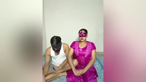 Video of a woman in a vibrant purple saree and a matching mask, seated next to a man in a white tank top and beige shorts, both facing forward in a minimalist room.