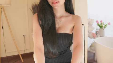 Video of a slender woman with long black hair, wearing a black, ribbed strapless dress, in a bright, modern bathroom with white walls, a freestanding bathtub, and a tall floor lamp.