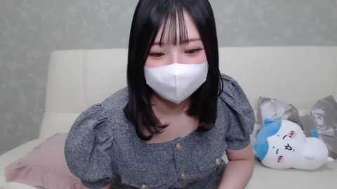Video of an East Asian woman with shoulder-length black hair, wearing a gray-patterned dress and white face mask, lying on a beige sofa, with a plush toy and a tissue box nearby.