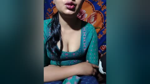 Video of a young woman with light brown skin, wearing a teal, low-cut top with pink floral embroidery, sitting on a blue sofa. The background features a colorful, ornate tapestry with orange and blue floral patterns.