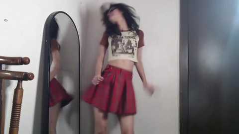 Video of a young woman with long black hair, wearing a cropped graphic tee and a red pleated skirt, dancing in front of a mirror, with a wooden chair in the background.
