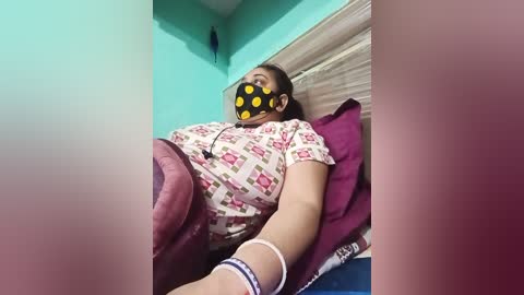 A video shows a woman lying on a bed, wearing a black mask with yellow dots, a patterned shirt, and a pink blanket. The room has teal walls and wooden accents.
