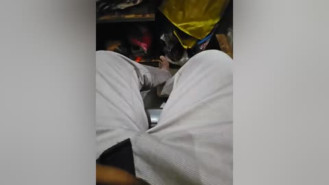 A video of a person lying on a bed with their legs spread, wearing light-colored pajamas. The room is cluttered with a yellow bag and various items in the background.