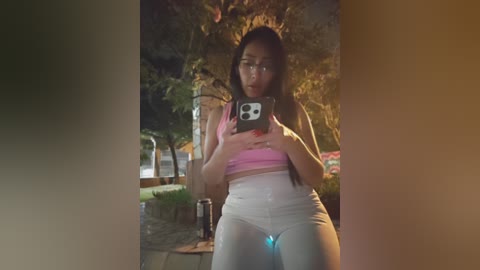 Video of an Asian woman with long black hair, wearing glasses, a pink crop top, and tight white pants, taking a selfie at night, surrounded by greenery.