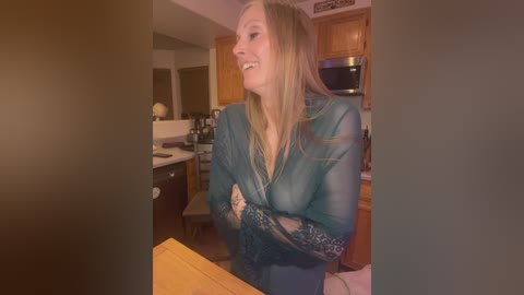 Video of a smiling, light-skinned woman with long blonde hair, wearing a sheer teal lace robe, in a cozy, dimly lit kitchen with wooden cabinets and a microwave.