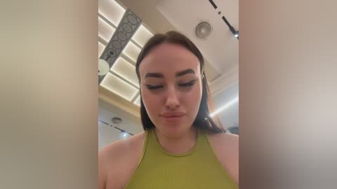 Video of a young woman with long brown hair, wearing a lime green ribbed tank top, standing in a modern room with white walls and a circular ceiling light.