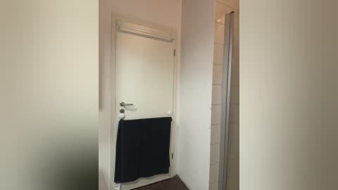 A video of a minimalist, modern bathroom featuring a closed white door with a black towel hanging on a hook. The walls are off-white, and a tiled shower is partially visible in the background.