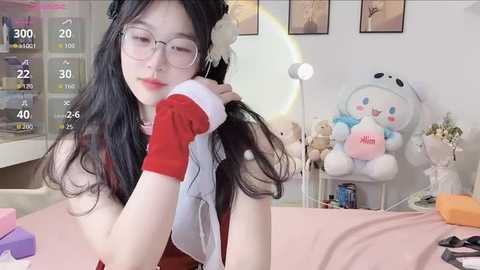 A video of an Asian woman with long black hair, wearing glasses, red top, and white gloves, adjusting her hair in a cozy room with a teddy bear and framed art on the wall.