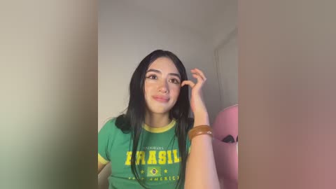 Video of a young woman with long black hair, wearing a green Brazil soccer jersey, touching her face. She has a light skin tone and is indoors with a blurred background.