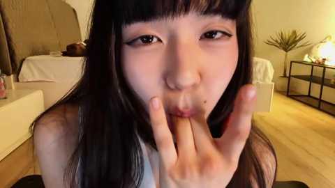 Video of an East Asian woman with long black hair, fair skin, and blunt bangs, making a peace sign with her hand. She is indoors, in a warmly lit bedroom with a white bed, nightstand, and potted plant.
