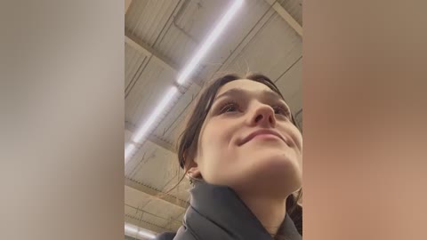Video of a young woman with fair skin and dark hair, wearing a grey jacket, looking up with a slight smile, against a beige ceiling with fluorescent lights.