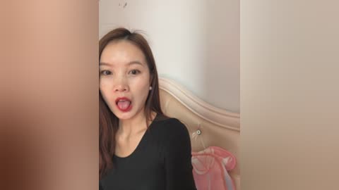 Media: A video of an Asian woman with long brown hair and fair skin, wearing a black top, appears shocked with her mouth open, surrounded by blurred beige walls.