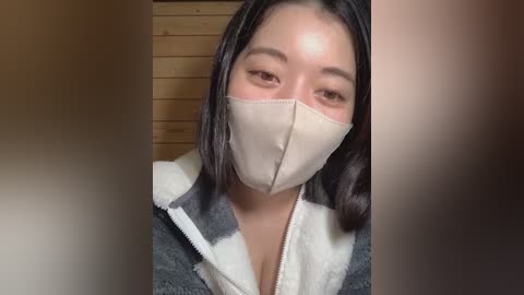 A video of an East Asian woman with shoulder-length dark hair, wearing a beige surgical mask, gray hoodie, and a white fleece lining, standing indoors against a wooden background.
