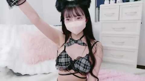 Video of an East Asian woman with long black hair and black cat ears, wearing a black plaid bralette, face mask, and black gloves, posing in a white bedroom with pastel accents.