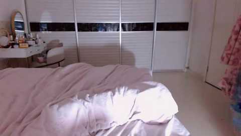 Video of a dimly lit, cozy bedroom with a white bed, light pink duvet, white dresser with mirror, white blinds, and a pink curtain on the right.